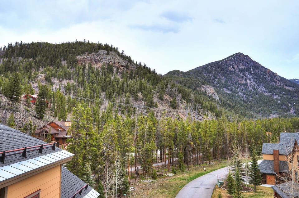 Mountainous landscape with green forested hills, houses with sloped roofs in the foreground, and a winding road. Clear sky with minimal cloud cover.