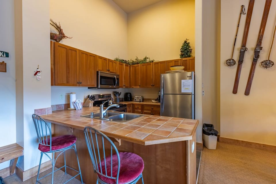 A kitchen with wooden cabinetry, stainless steel appliances, a tiled countertop island with two barstools, and vintage wooden skis mounted on the wall.
