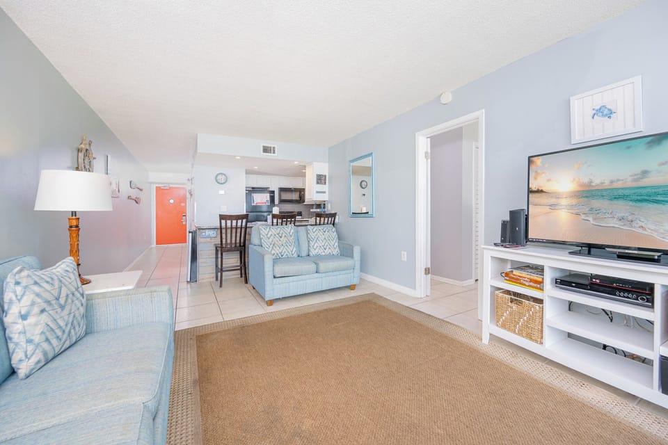 Cozy living area featuring cozy blue seating, a large TV, and a welcoming view into the modern kitchen