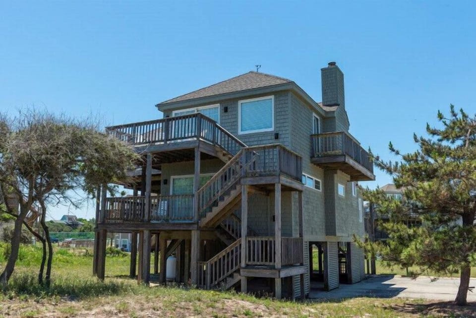 Coastal beach house elevated on stilts with wraparound decks offering panoramic views, nestled among windswept trees in a serene natural setting.