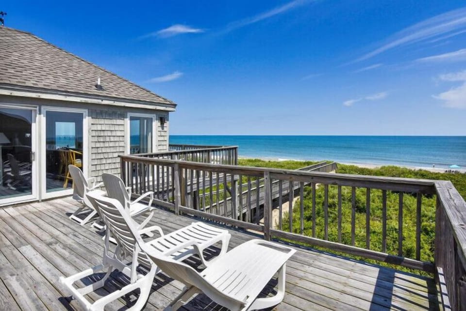 Relax on your private oceanfront deck, where white lounge chairs invite you to unwind while taking in sweeping views of turquoise waters and coastal greenery under endless blue skies.