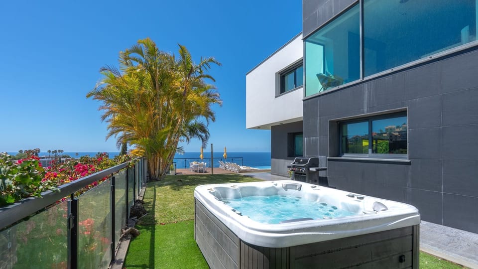 Private garden terrace features a bubbling hot tub beside a sleek modern villa, framed by palms and ocean views. Glass railings, manicured lawn, and warm sunlight invite effortless resort-style relaxation.
#HotTub #LuxuryVilla