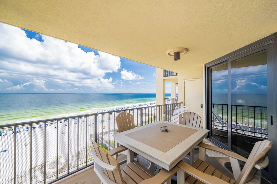 The balcony features a dining table and chairs with a breathtaking view of the beach and vibrant blue waters