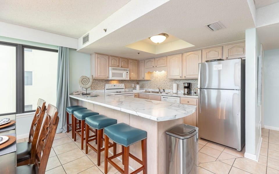 This kitchen features white and stainless appliances, a spacious countertop, and a cozy dining area with teal bar stools