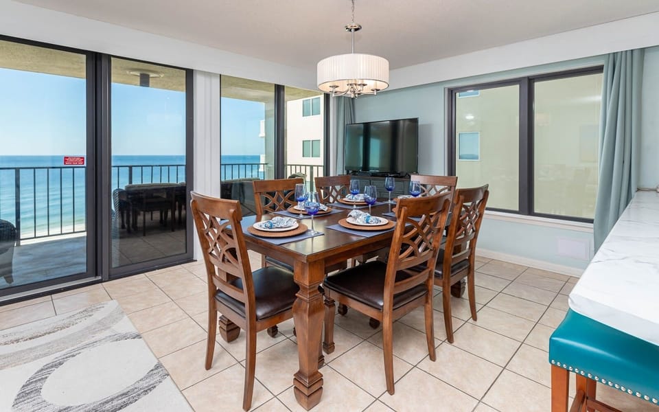 This dining area features a stylish wooden table set for six, complemented by a large TV and a breathtaking beach view
