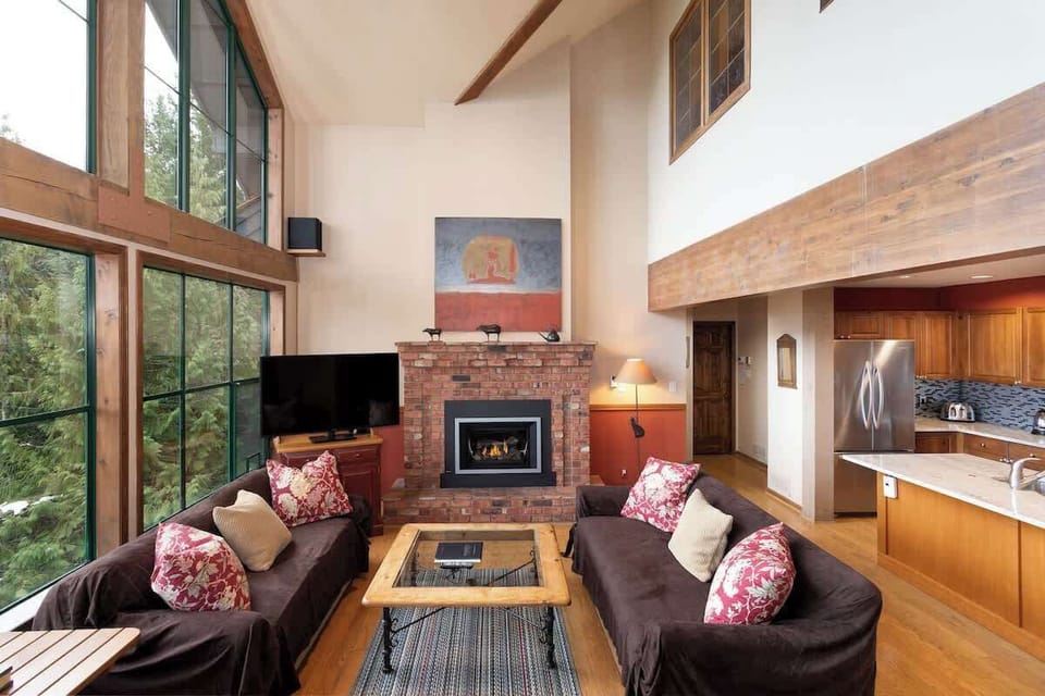 - Spacious living area with towering windows and forest backdrop  
- Brick fireplace and plush sofas create a warm mountain retreat  
- Open layout connects seamlessly to dining and modern kitchen