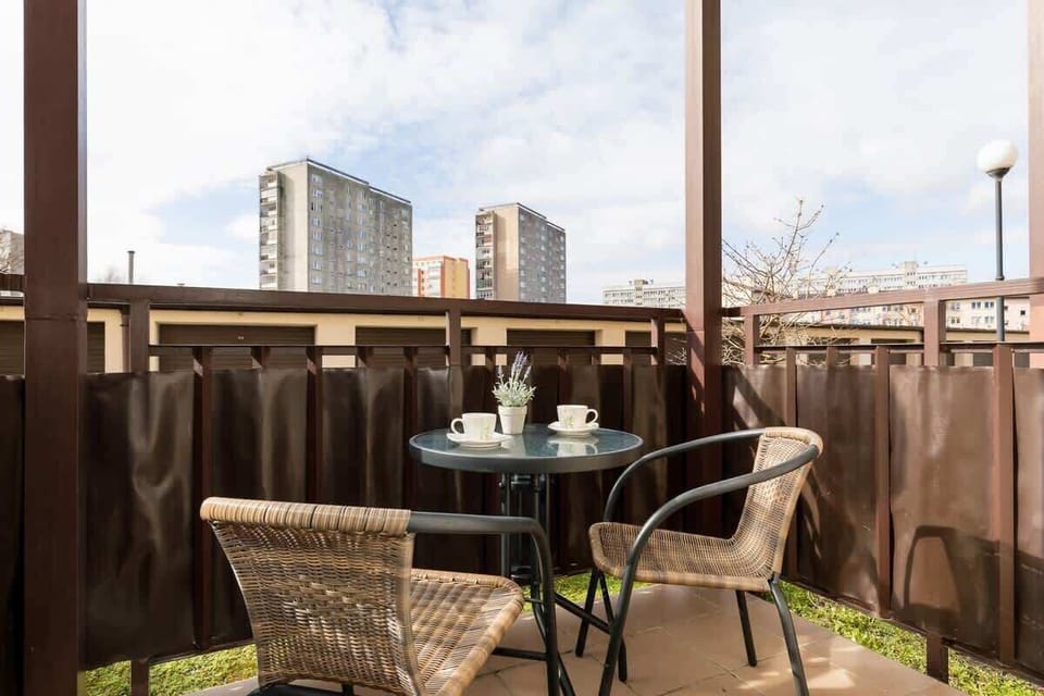 An outdoor balcony with seating and a small table, overlooking an urban landscape. The balcony has a metal railing and provides an outdoor relaxation space.