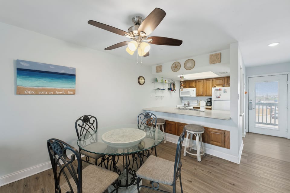 Gather around this cozy dining area, where coastal vibes and beach time decor set the tone for your getaway, complete with an open kitchen and balcony access nearby.