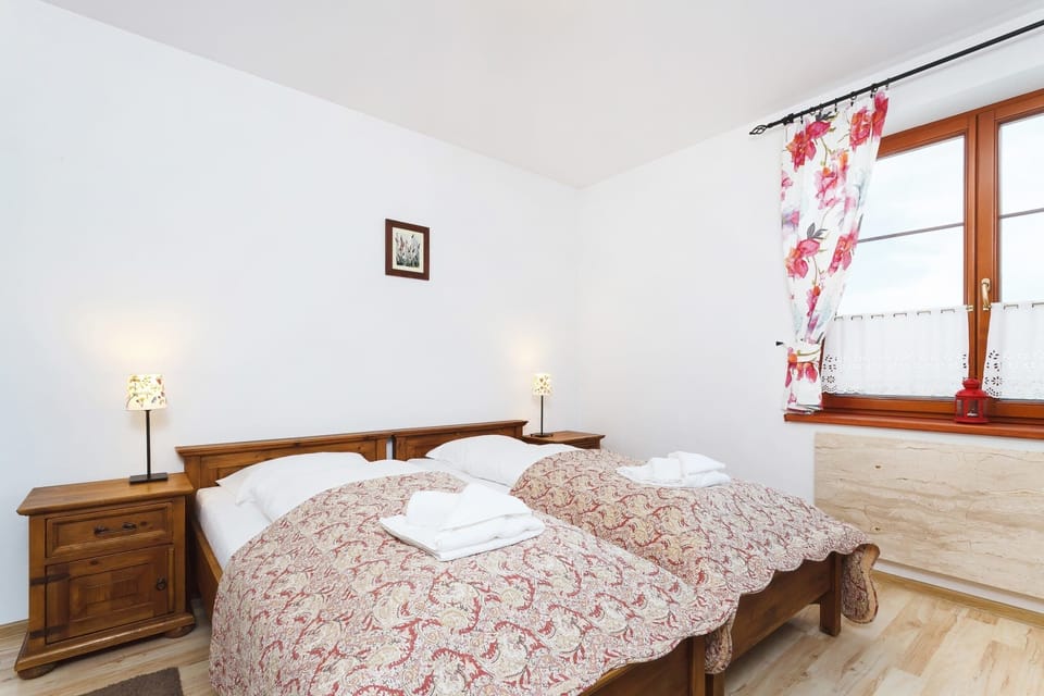  A simple bedroom with a single beds covered in floral bedding, a wooden nightstand with a lamp, and a small window offering natural light.