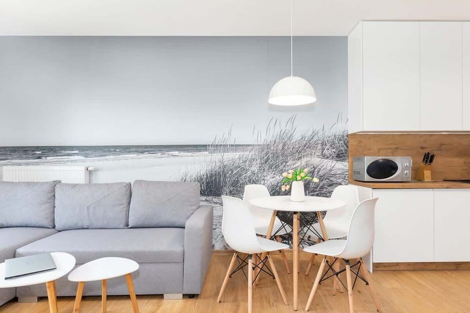 Bright living room with a grey sofa, round dining table, and an open kitchenette with white cabinets and wood accents.