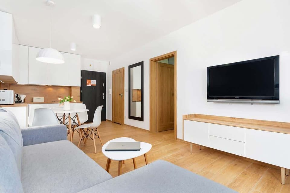 A bright living room with a small round dining table and chairs, a grey sofa, and a modern kitchen area in the background.