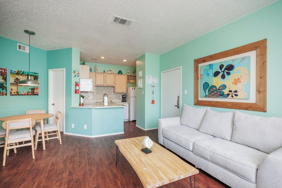 Relax in your bright, coastal-themed living space with cheerful turquoise walls, comfortable seating, and an open kitchen perfect for preparing meals together.