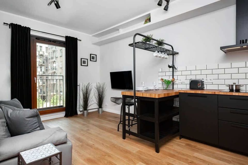 A stylish living room with an open-plan kitchenette, featuring a comfortable sofa and a dining area. The modern design includes sleek black cabinets in the kitchen area, blending seamlessly with the living space.
