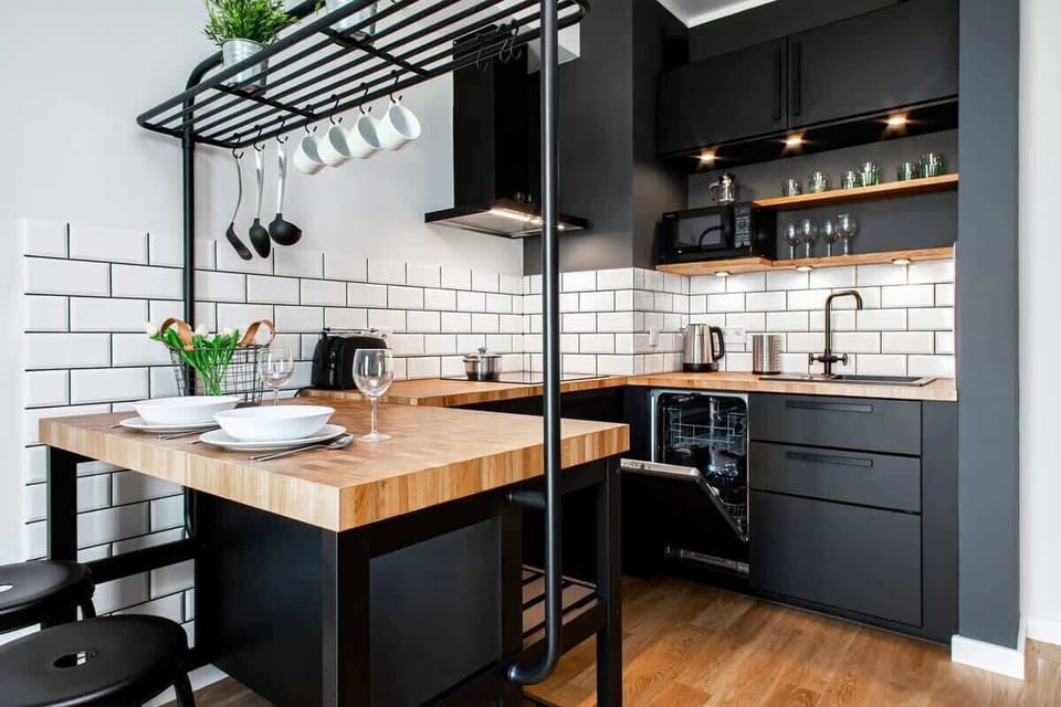 A sleek black kitchen with white tile backsplash, open shelving, and a wooden countertop. There are industrial pendant lights hanging above the kitchen island.