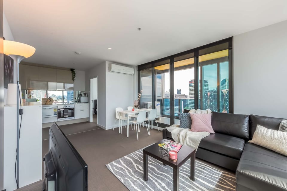 An open-plan space features a living area furnished with an L-sofa with a fold-out sofa bed, floor-to-ceiling windows, a wooden coffee table, and a wide-screen TV.