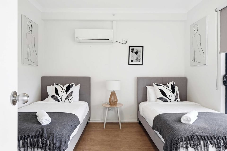 Relax in this twin bedroom featuring two plush single beds, a split-system air conditioner, bedside table with a stylish lamp, blackout roller blinds, and contemporary wall art—designed for a comfortable and restful stay.