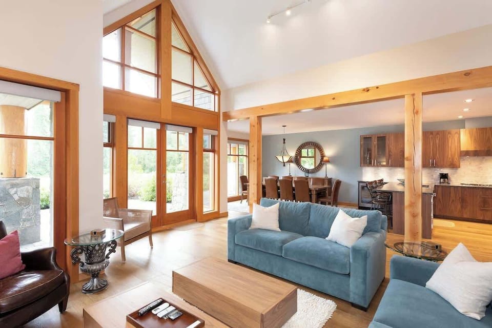 - Bright open living room with vaulted ceilings and tall windows framing lush views  
- Soft-toned furniture and warm wood details create a welcoming mountain vibe  
- Spacious flow connecting lounge, dining, and kitchen areas for easy gatherings  
