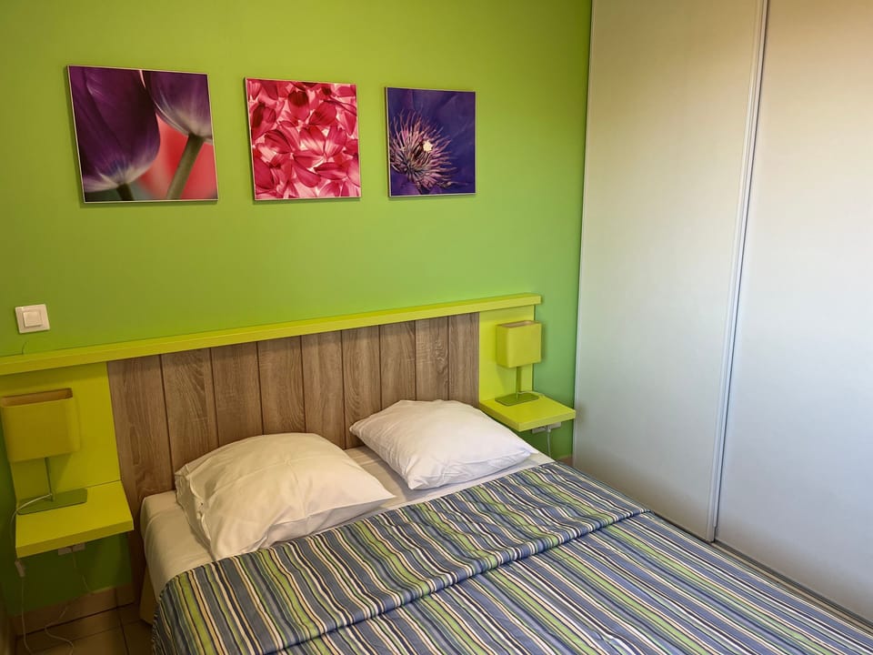 The master bedroom features a double bed with a wood-panel headboard set against fresh green walls and floral wall art.