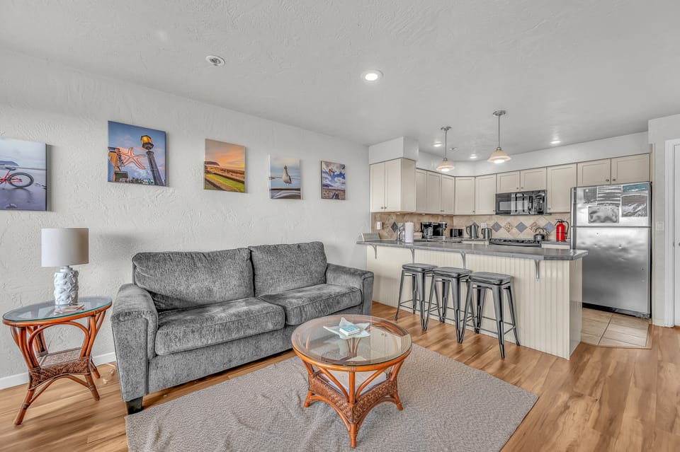 Relax in your stylish open-plan living space, where modern comfort meets coastal charm with warm wood floors and a fully equipped kitchen.
