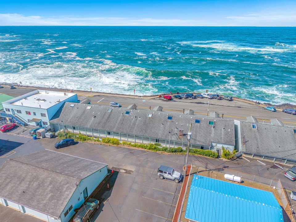 Aerial view of beachfront property featuring swimming pool and parking areas, with stunning ocean waves just steps from the building.