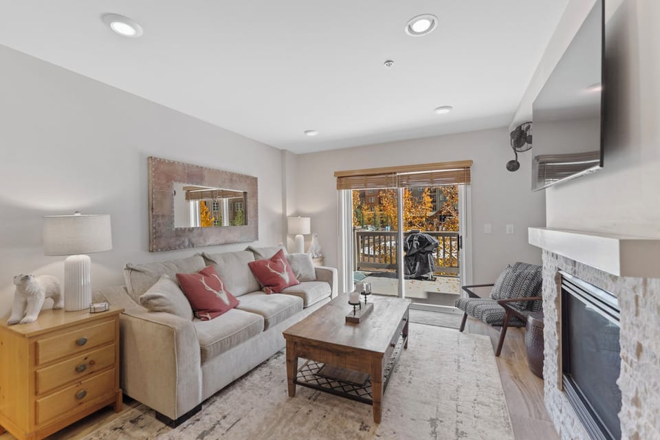 Relax in your cozy living room with plush seating, warm fireplace, and peaceful balcony views of autumn foliage.