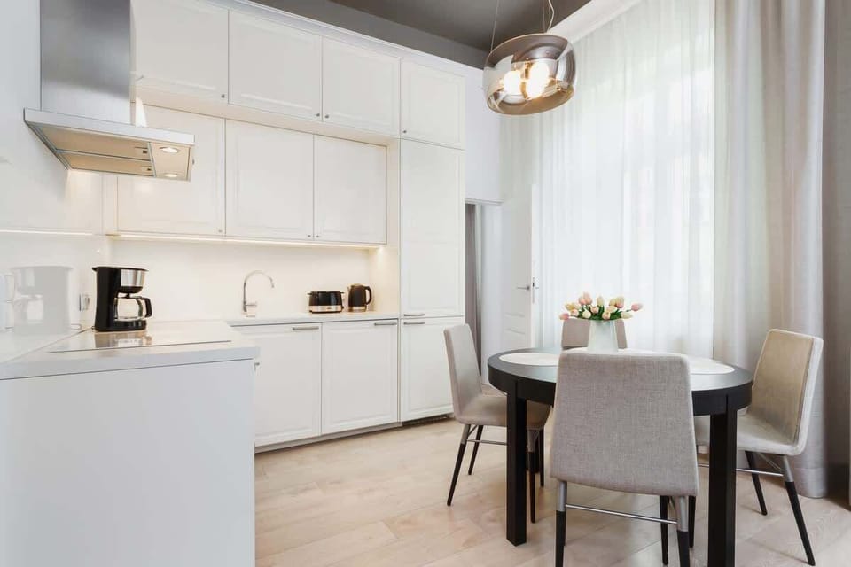 A wider view of a kitchen combined with a small dining area. It features white cabinets, a dining table, and a few decorative plants.