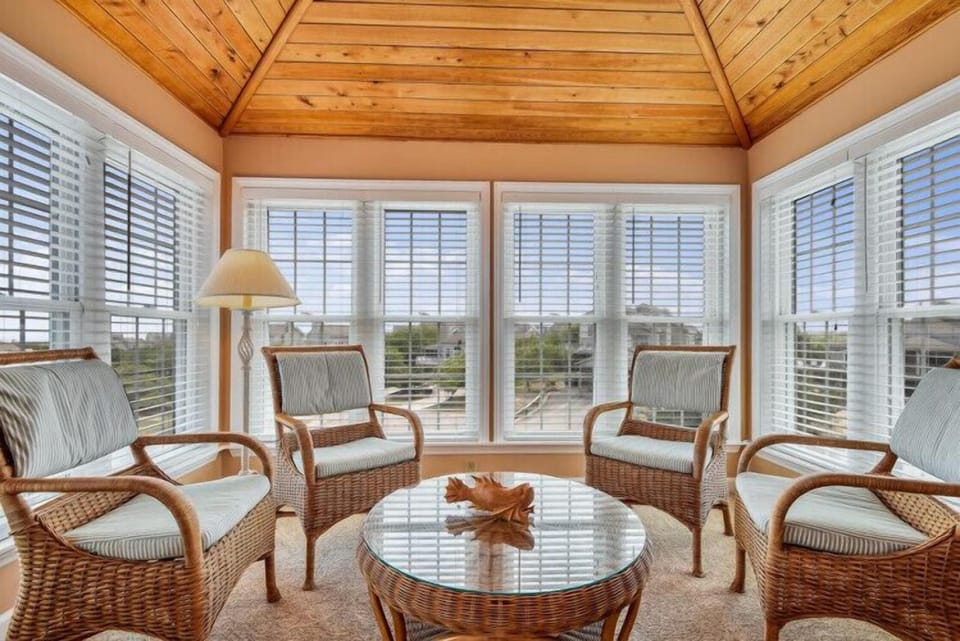 Relax in this bright sunroom with wicker seating, wooden vaulted ceilings, and panoramic windows framing serene outdoor views—a peaceful space to unwind any time of day.