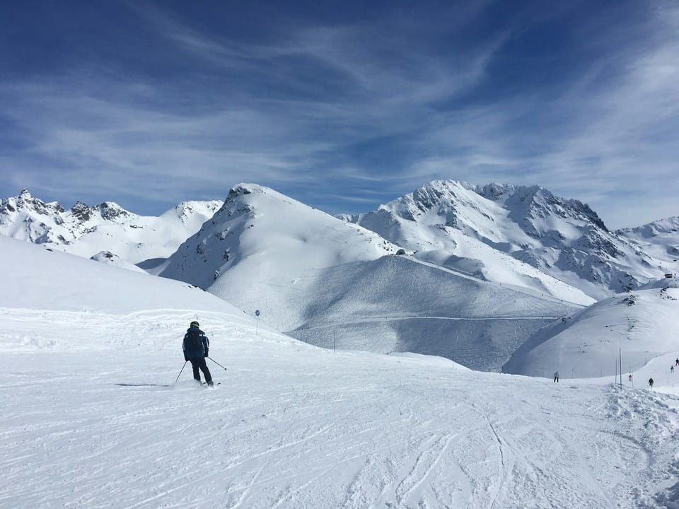 A lone skier carves down a wide Les Menuires piste beneath the dramatic high-altitude peaks of the Les 3 Vallées, one of the world's largest ski areas.