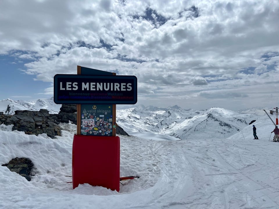 The Les Menuires resort sign marks the gateway to the slopes, with the vast snow-covered peaks of the Les 3 Vallées ski area stretching behind.