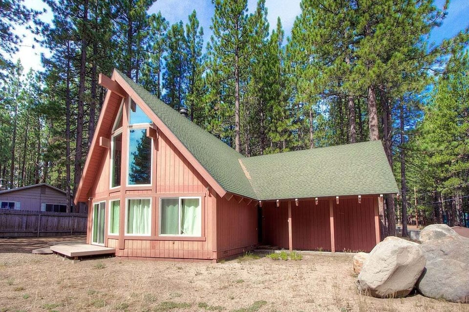 Exterior with A-frame house, large windows, deck, chimney, trees, fence, and boulders.