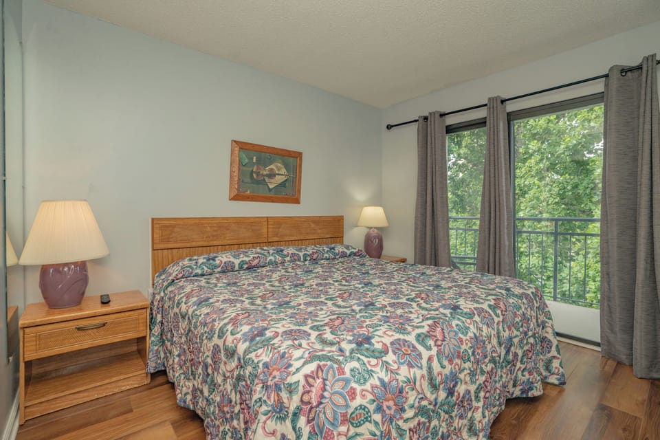 Unwind in your peaceful retreat with a comfortable bed framed by warm wood accents and a calming view of lush greenery through your private balcony door.