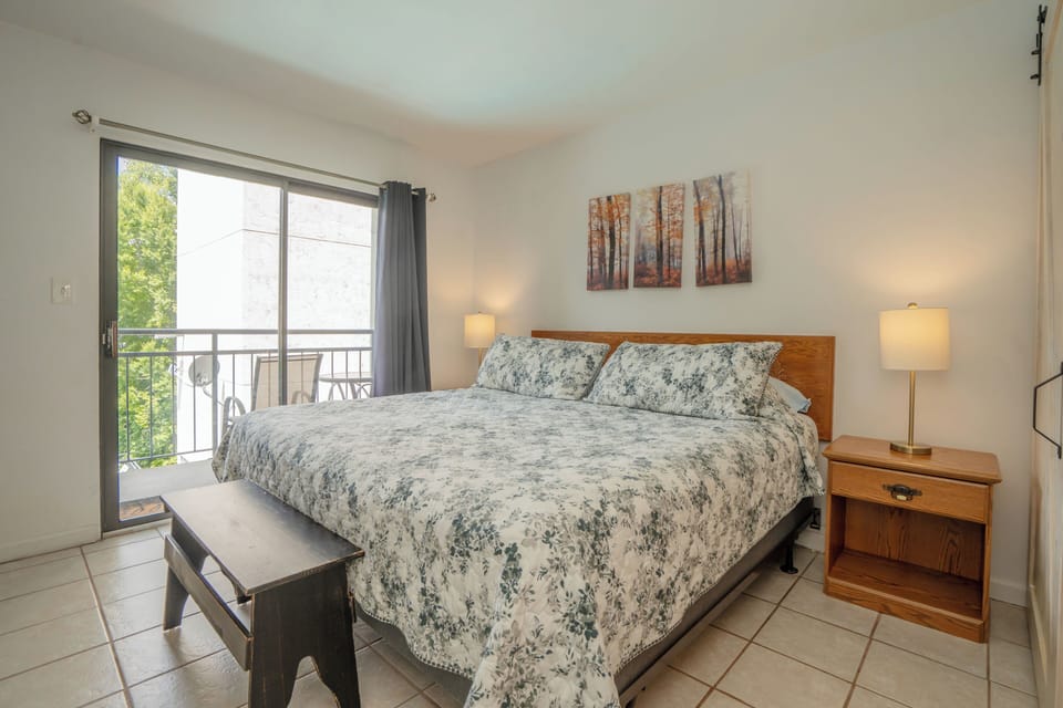 Relax in your cozy bedroom with soft bedding and natural light streaming through the balcony door. Step outside for a breath of fresh air or unwind with the peaceful wooded scene above the bed.