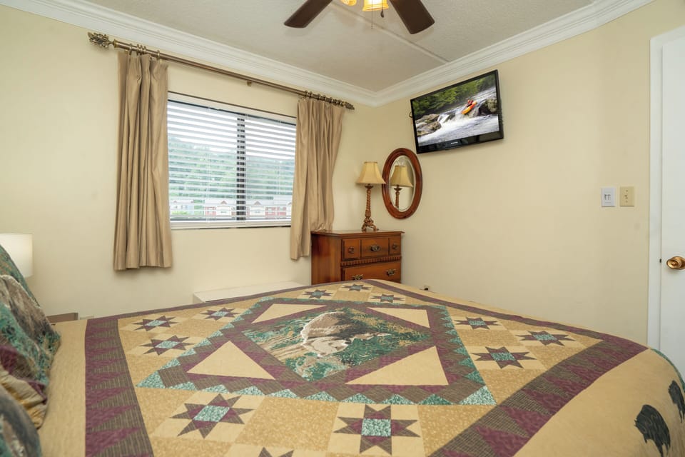 Your bedroom features a handsome quilt-topped bed and warm wooden furnishings, lit by soft natural light through the window and the gentle glow of a ceiling fan above.