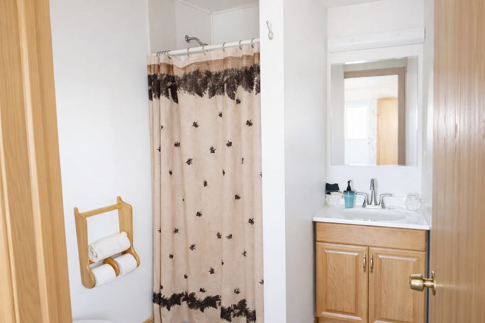 Cozy country-style bathroom with all the essentials — featuring a warm pine-themed shower curtain, soft bath mat, and rustic wood accents for a simple, homey feel.