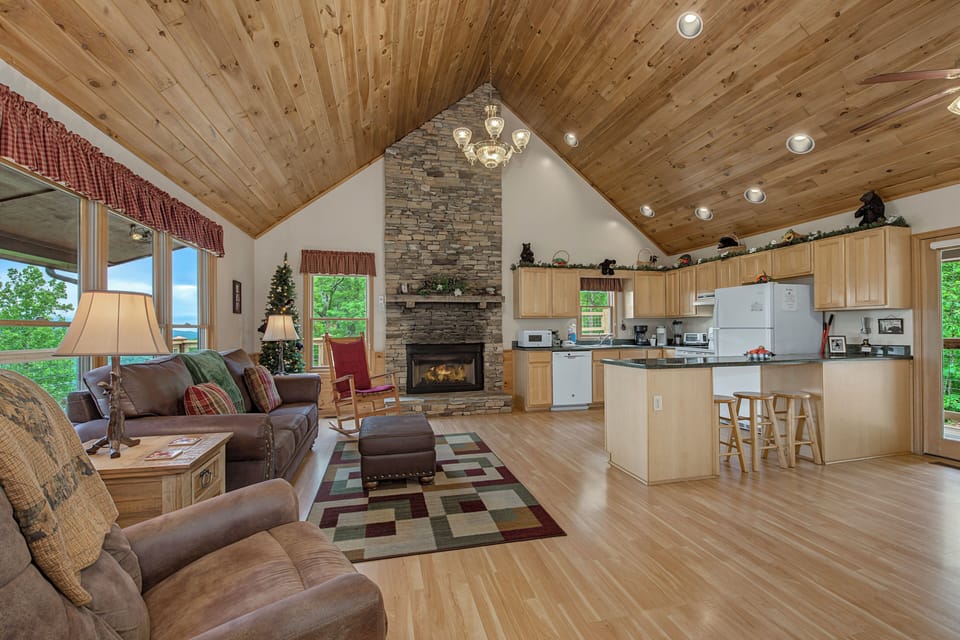Relax in your open-concept cabin featuring vaulted wood ceilings, a cozy stone fireplace, plush seating, and a fully equipped kitchen—perfect for mountain getaways.