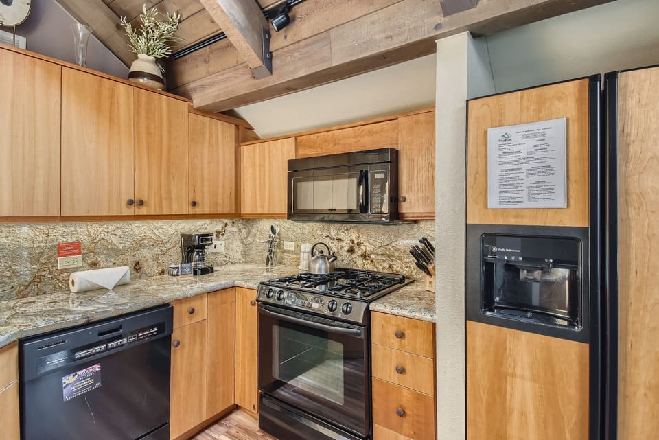 A kitchen featuring wooden cabinets, a gas stove, a microwave, a dishwasher, and a black refrigerator. Granite countertops and a mounted plant contribute to the decor.