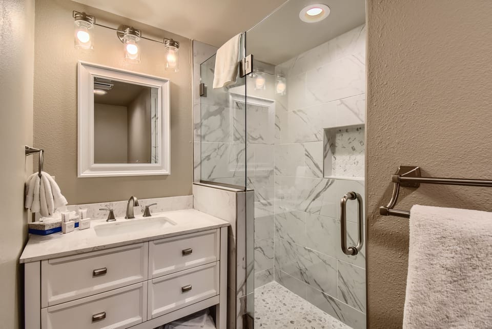 A modern bathroom with a white vanity, marble countertop, rectangular mirror, glass shower enclosure, and marble-tiled shower walls and floor.