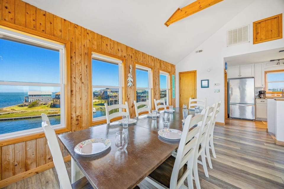 Picture yourself savoring morning coffee at this sunlit dining table, where floor-to-ceiling windows frame stunning waterfront views. Warm wood accents and vaulted ceilings create a welcoming space for memorable meals together.