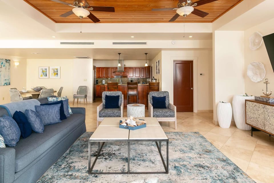 Living area with navy cushions on gray sofa, patterned armchairs, tiled floor, and wood-paneled ceiling fans.