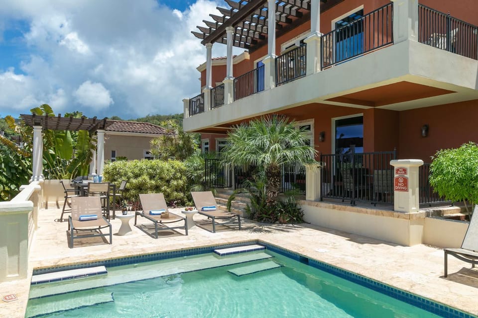 Sunlit pool deck with beige loungers, side tables, potted palms, and a shaded dining area under a wooden pergola.
