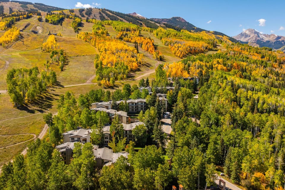 Aerial view of the property nestled in a spectacular mountain setting, surrounded by golden aspens and evergreens with ski slopes and dramatic peaks rising in the distance.