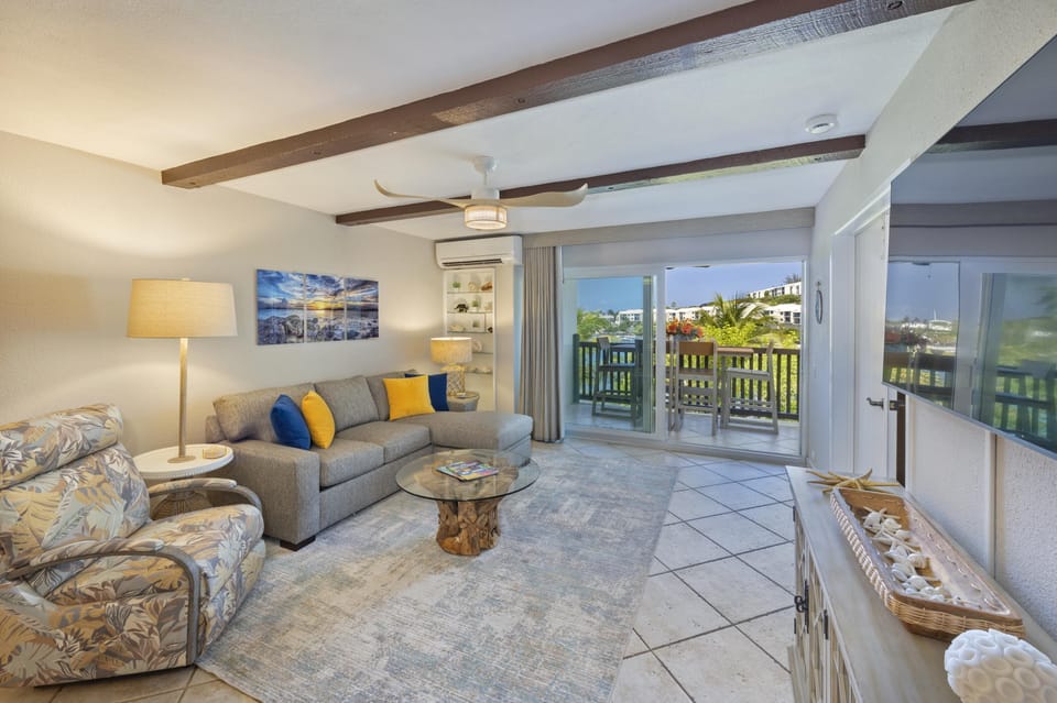 Living room with sectional sleeper sofa, floral recliner, glass coffee table with driftwood base, ceiling fan, and sliding doors to balcony.