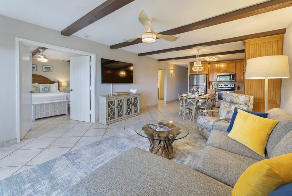 Open-concept living room and dining area with tiled floors, exposed ceiling beams, and a driftwood base glass coffee table.