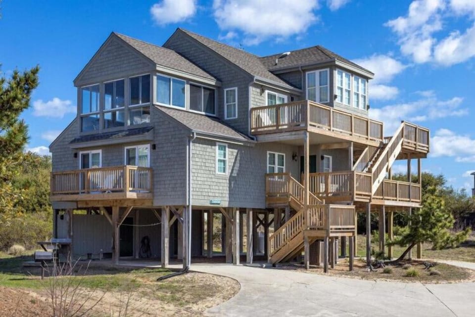A classic coastal beach house elevated on stilts, featuring multiple levels of wooden decks and balconies overlooking the natural landscape, offering a perfect seaside retreat.