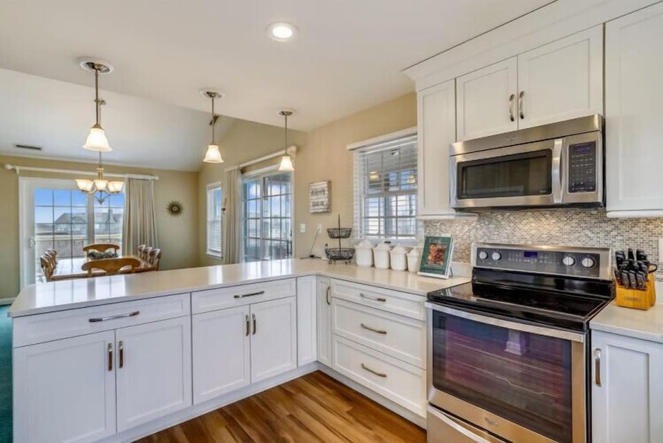 Your bright, fully equipped kitchen invites culinary adventures with gleaming white countertops, modern appliances, and rich wood floors that flow into a sunny dining nook with serene outdoor views.
