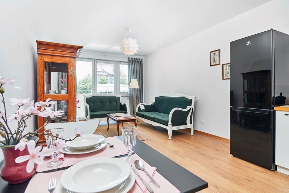 The dining table is elegantly set with plates, glasses, and a vase of flowers, adding charm to the space. In the background, the living room features a green sofa and a wooden TV cabinet, creating a cozy atmosphere