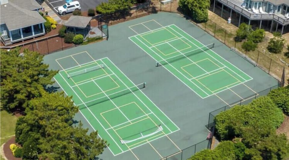 Aerial view of community tennis courts with multiple well-maintained surfaces, surrounded by landscaping and neighboring homes, available for guest use during your stay.