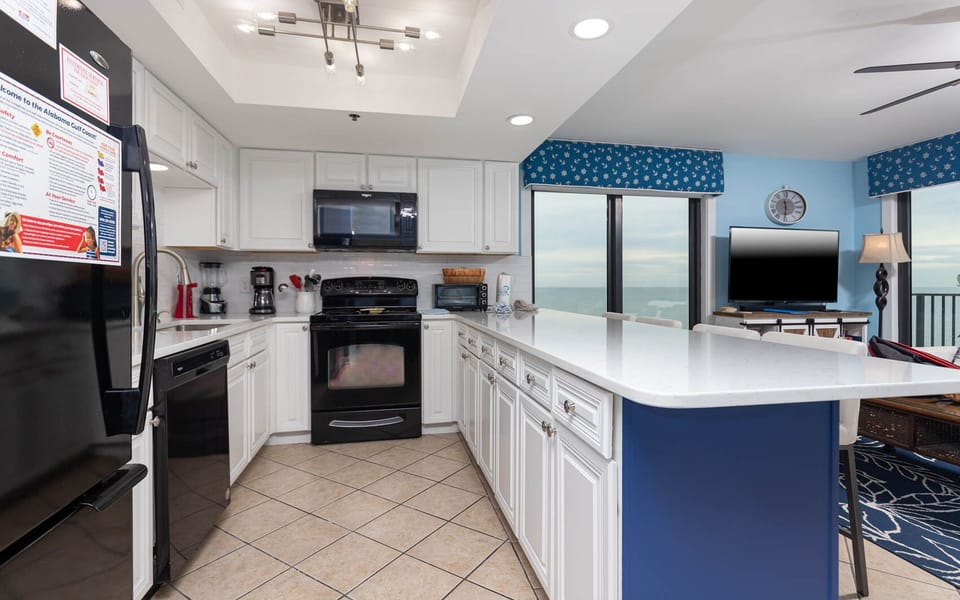 This kitchen features sleek black appliances, a coffee maker, and a beautiful view of the beach from the dining area