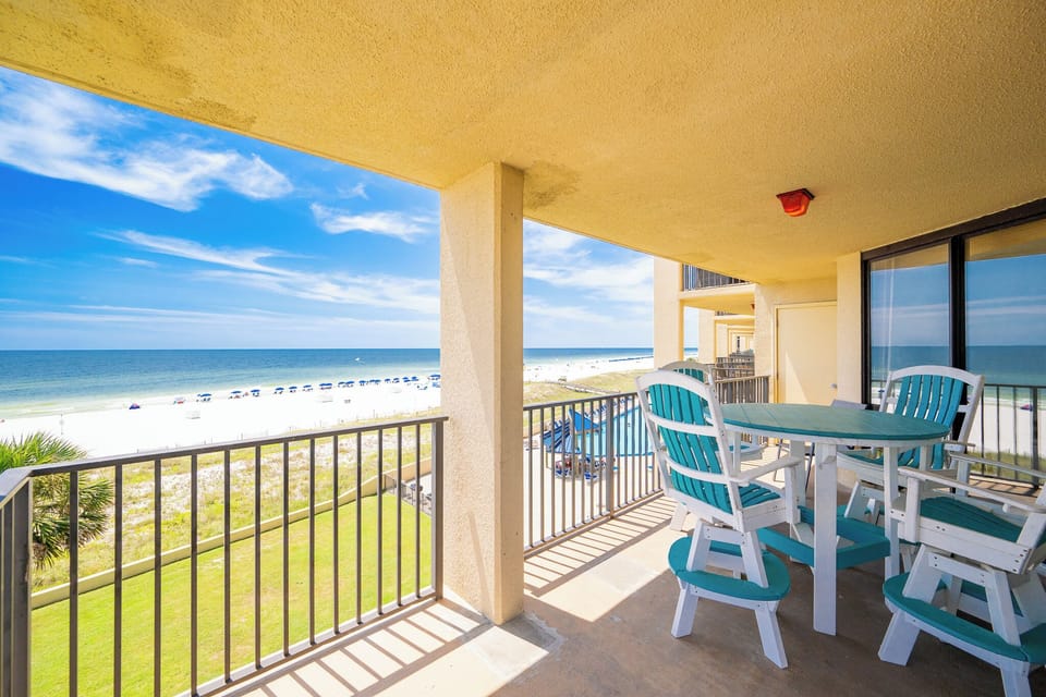 The balcony features a round table with teal chairs, offering a direct view of the beach and the inviting blue waters