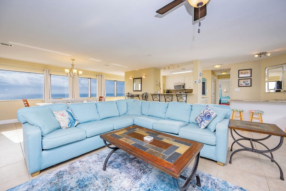 The living area features a blue sectional sofa, a stylish coffee table, and stunning beach views through large windows